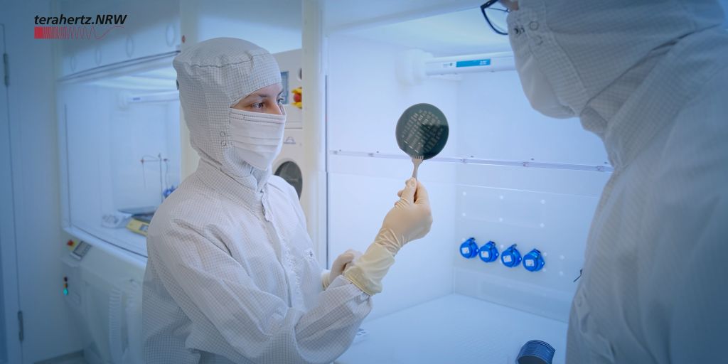Zwei Personen in Reinraumanzügen arbeiten in einem Labor. Eine Person hält eine runde Silizium-Waferplatte mit Pinzette ins Licht. Im Hintergrund sind Reinraumgeräte zu sehen. Oben links ist das Logo von terahertz.NRW.