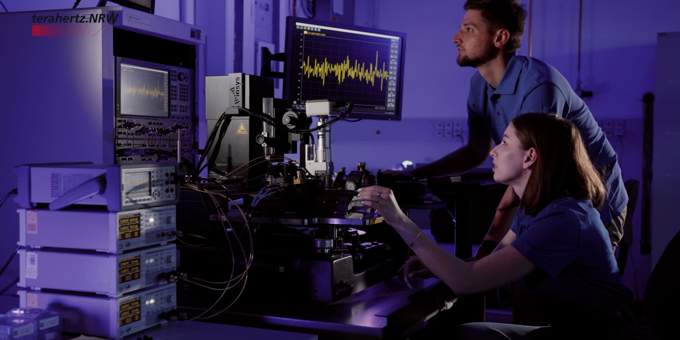 Zwei Forschende in einem Labor bedienen ein komplexes Messsystem für Terahertzsensorik mit Oszilloskop und mehreren Bildschirmen. Auf den Monitoren sind Wellendiagramme zu sehen. Two researchers in a laboratory operate a complex measurement system for terahertz sensing with an oscilloscope and multiple screens. Waveform diagrams are visible on the monitors.