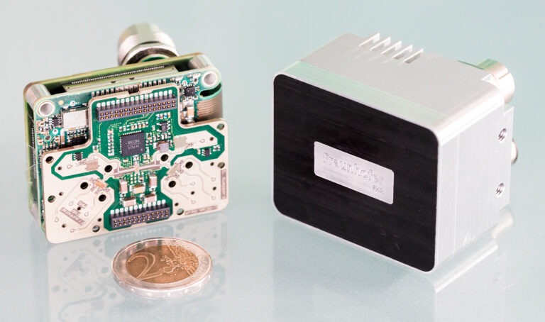 Kompakte Radarsensor-Einheit neben einer 2-Euro-Münze zum Größenvergleich, entwickelt von Fraunhofer FHR für Terahertzsensorik. Compact radar system for terahertz sensing placed next to a coin for size reference.