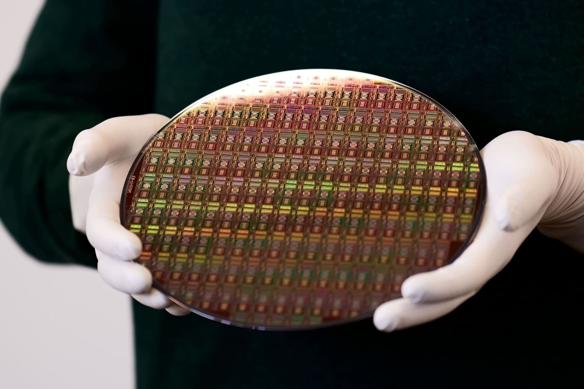 Eine Person mit weißen Handschuhen hält einen Siliziumwafer, auf dem zahlreiche Mikrochips für die Terahertzsensorik in regelmäßiger Anordnung zu sehen sind. Die Oberfläche des Wafers zeigt einen schillernden Farbeffekt durch Lichtreflexionen. A person wearing white gloves holds a silicon wafer showing numerous microchips for terahertz sensing arranged in a regular pattern. The wafer surface displays an iridescent color effect from light reflections.