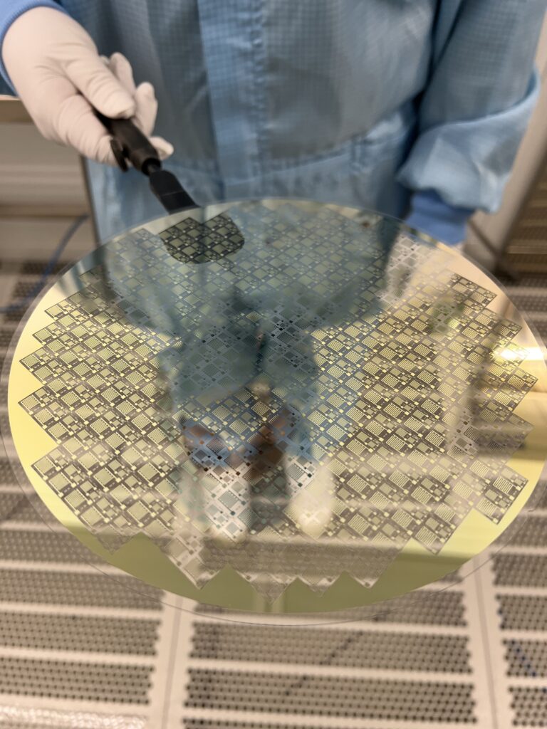 Eine Person in Reinraumkleidung hält eine teils goldene, teils transparent schimmernde Wafer-Scheibe zur Entwicklung von Terahertzsensorik. A person in cleanroom attire holds a wafer disc, partly golden and partly transparently shimmering, for terahertz sensing development.