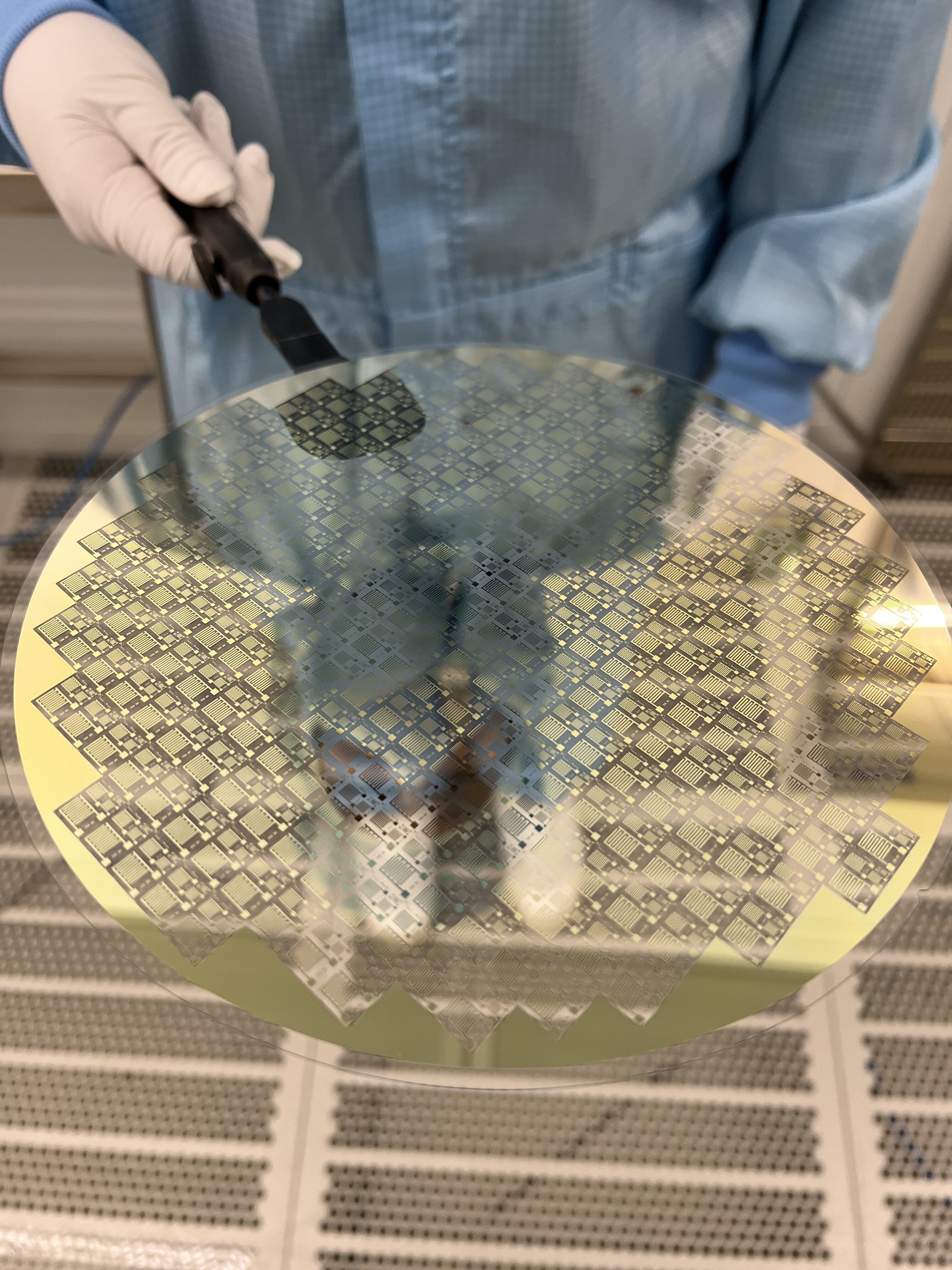 Eine Person in Reinraumkleidung hält eine teils goldene, teils transparent schimmernde Wafer-Scheibe zur Entwicklung von Terahertzsensorik. A person in cleanroom attire holds a wafer disc, partly golden and partly transparently shimmering, for terahertz sensing development.