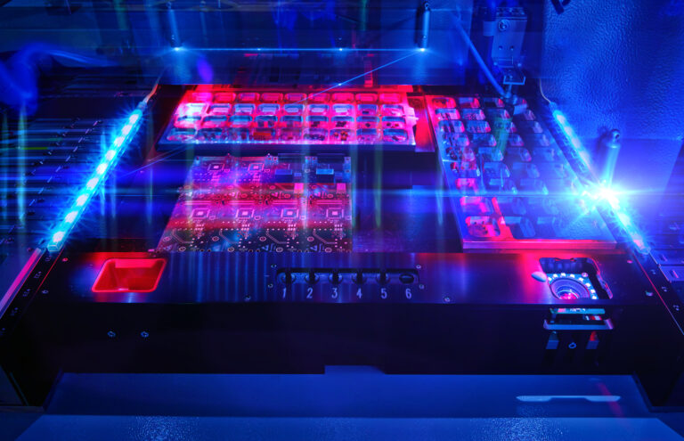 Ein Bestückungsautomat für Bauelemente, Chips und FPGA auf Platinen zur Entwicklung und Fertigung von Terahertzsensorik ist rot und blau beleuchtet. A pick-and-place machine for components, chips, and FPGAs on circuit boards for the development and production of terahertz sensing is illuminated in red and blue.