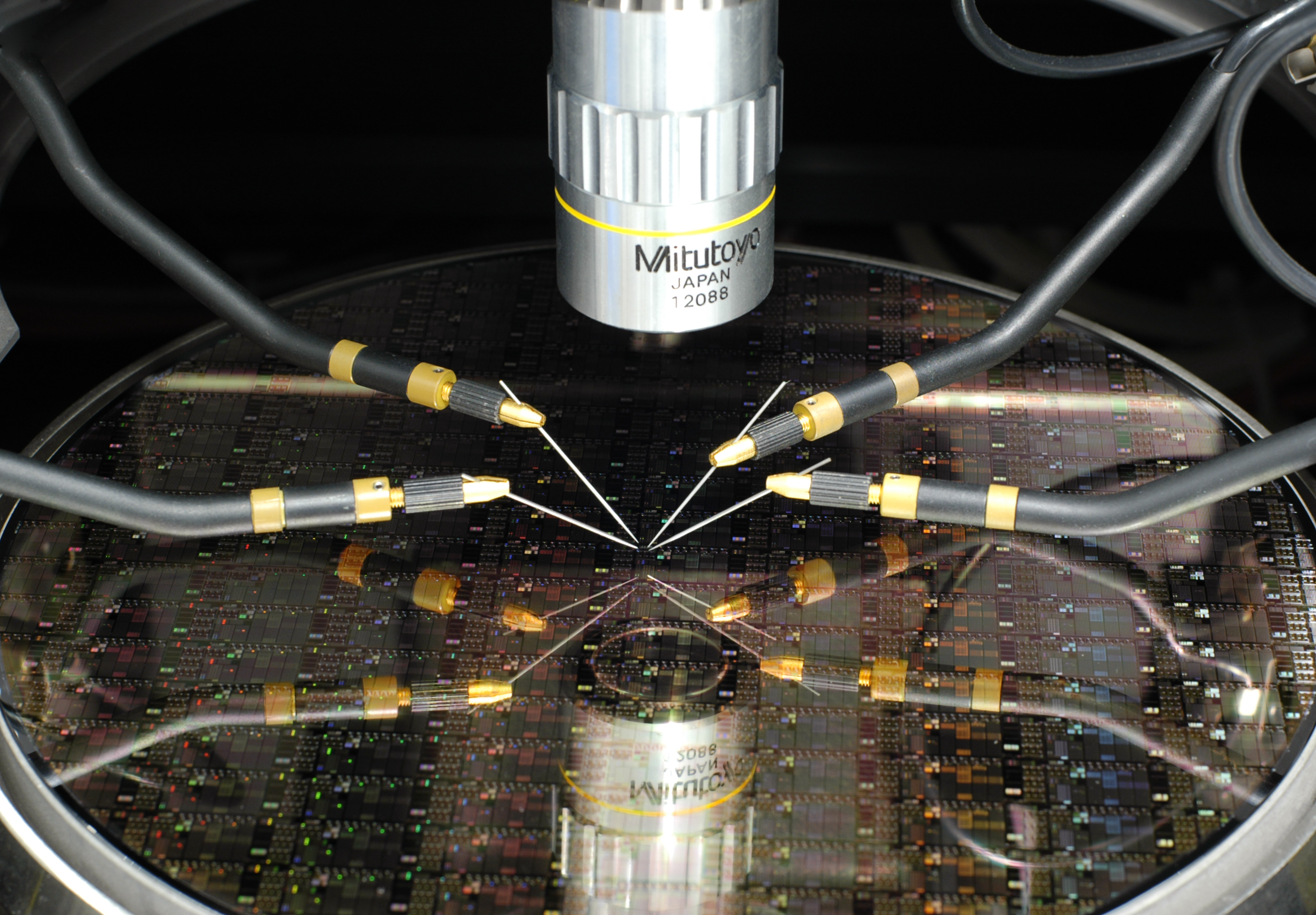 Halterungen mit sehr feinen Nadeln zur Charakterisierung von Schaltkreisen der Terahertzsensorik befinden sich über einem Wafer unter dem Mikroskop. Probes with very fine needles for characterizing integrated circuits in terahertz sensing, positioned over a wafer under a microscope.