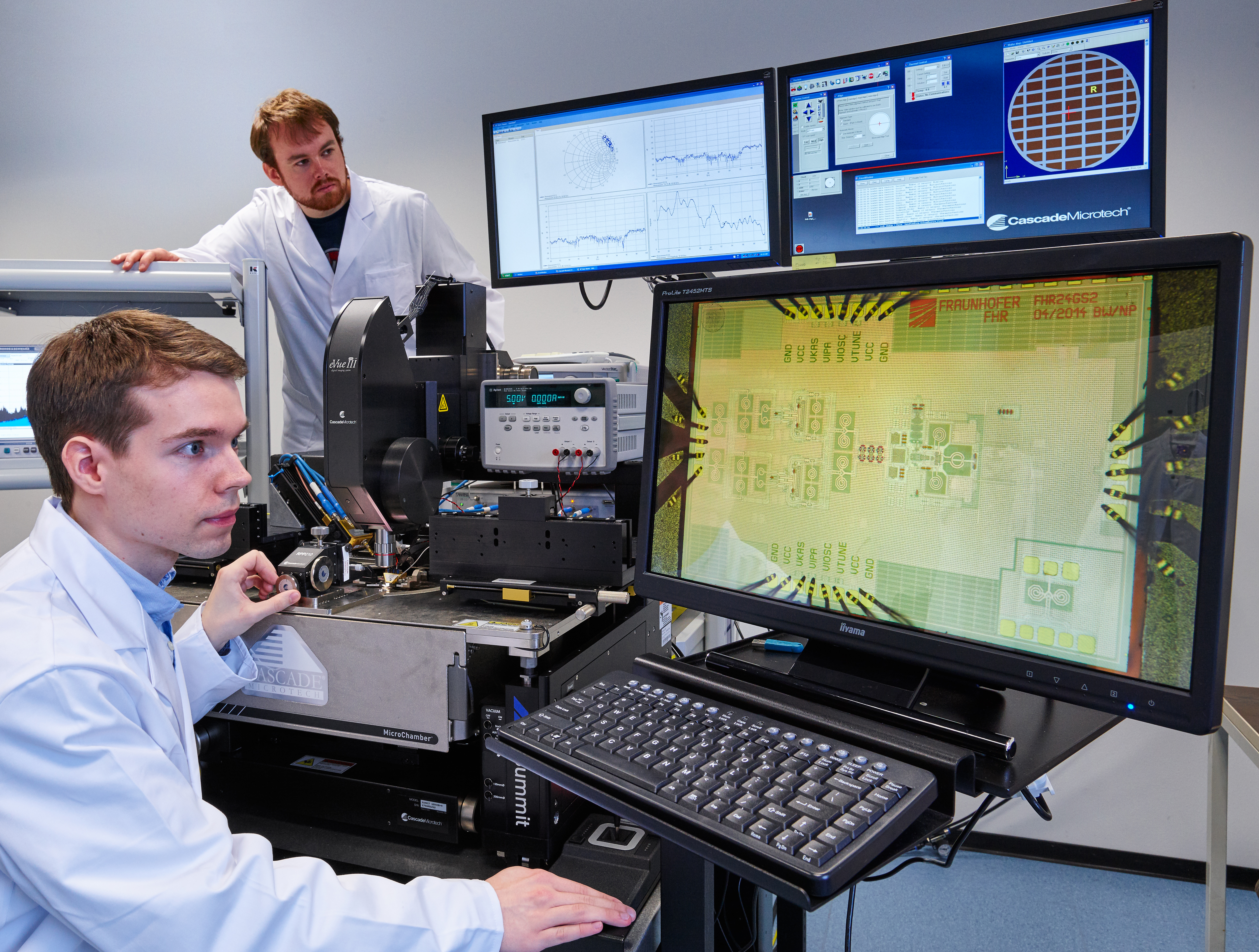 Zwei Wissenschaftler analysieren SiGe-Chips frü die Terahertzsensorik auf einem Wafer an einer On-Wafer-Messstelle und sehen die Ergebnisse auf Bildschirmen. Two scientists analyze SiGe chips for terahertz sensing on a wafer at an on-wafer measurement station, viewing the results on screens.